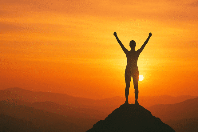 Silhouette of a confident woman standing on a mountaintop with arms raised at sunrise, symbolizing empowerment, self-worth, and confidence against a warm golden sky. Image is used for know your worth quotes
