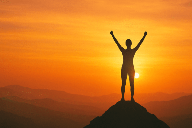 Silhouette of a confident woman standing on a mountaintop with arms raised at sunrise, symbolizing empowerment, self-worth, and confidence against a warm golden sky. Image is used for know your worth quotes